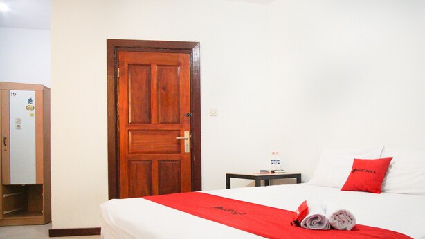 Habitación de hotel con puerta de madera, cama con ropa de cama blanca y roja, una mesita con una botella y un cojín rojo.