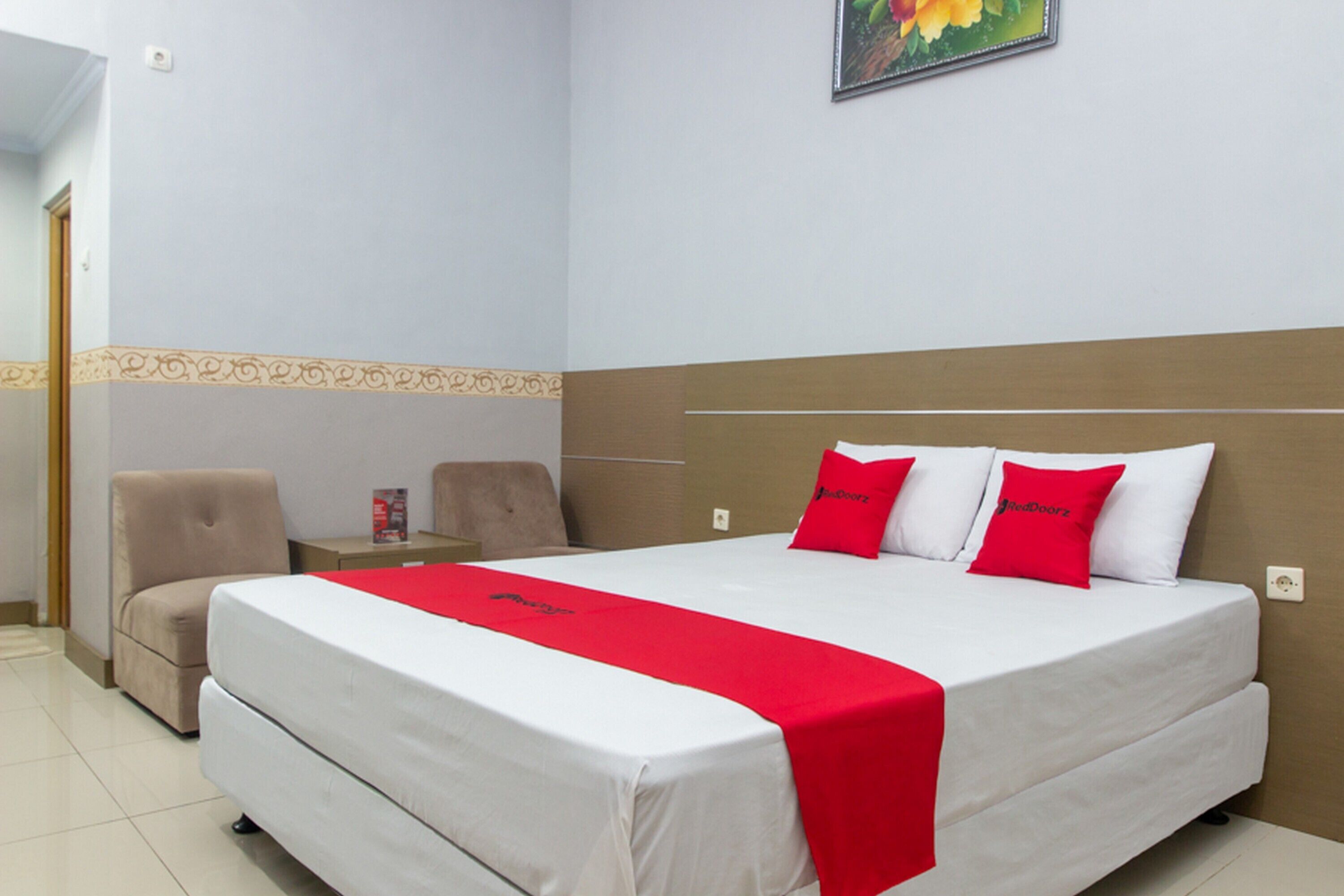 A hotel room with a large bed, two red pillows, a red bedspread, a beige armchair, and a nightstand with a red object on it.