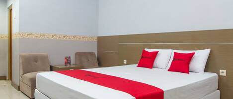 A hotel room with a large bed, two red pillows, a red bedspread, a beige armchair, and a nightstand with a red object on it.