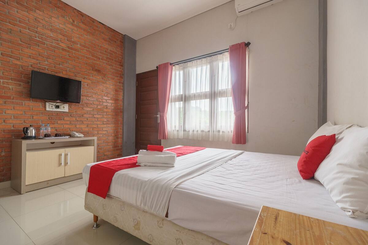 A hotel room with a brick wall, a flat-screen TV mounted above a wooden desk, a bed with white bedding and red accents, and a window with pink curtains.