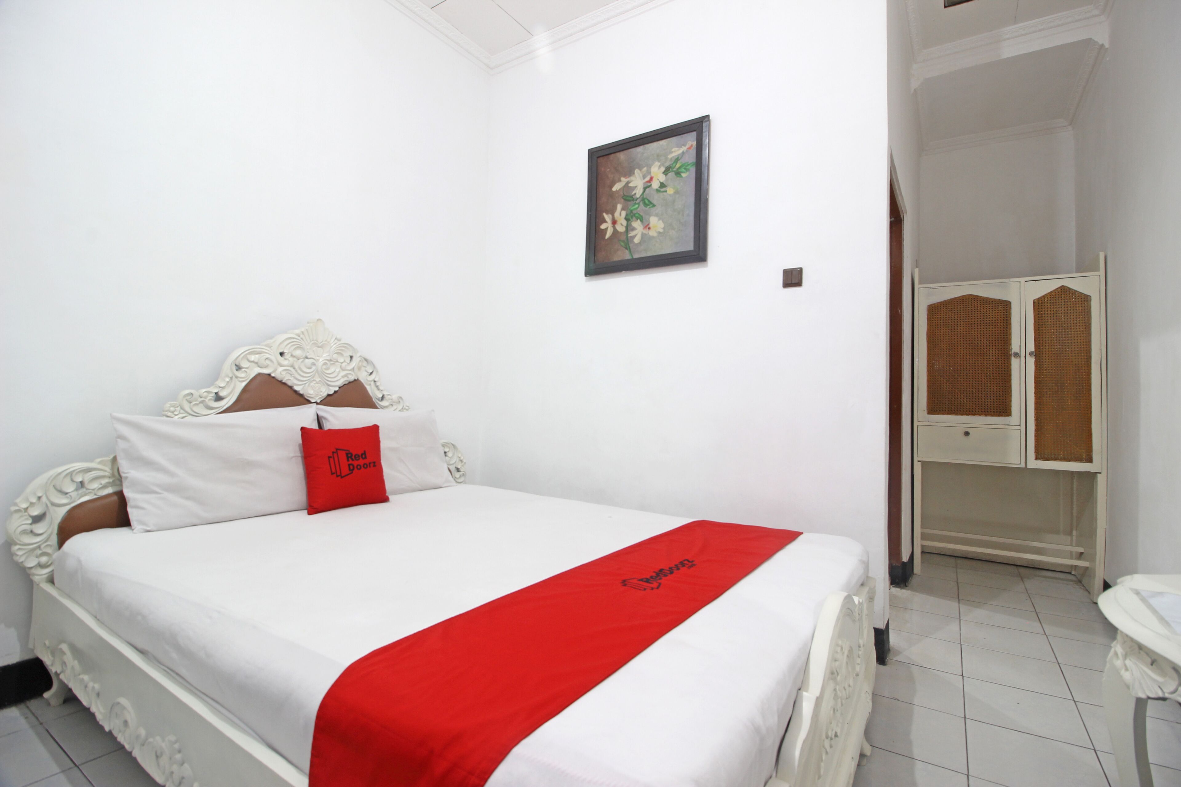 A white four-poster bed with a red throw, a framed floral painting on the wall, and a white wardrobe with a wicker door.