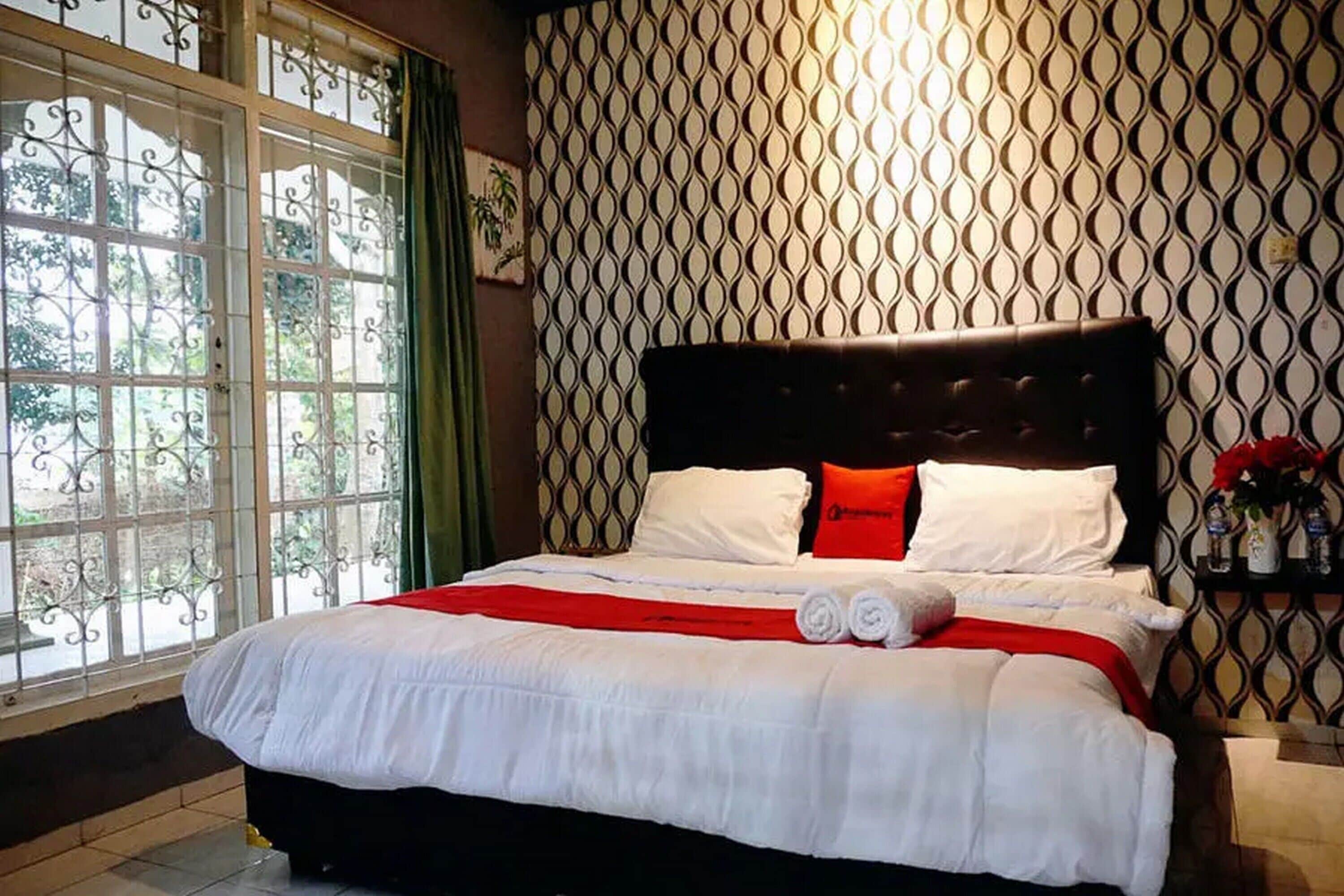 A neatly made bed with white bedding and red accents, a dark headboard, and a patterned wall behind it.