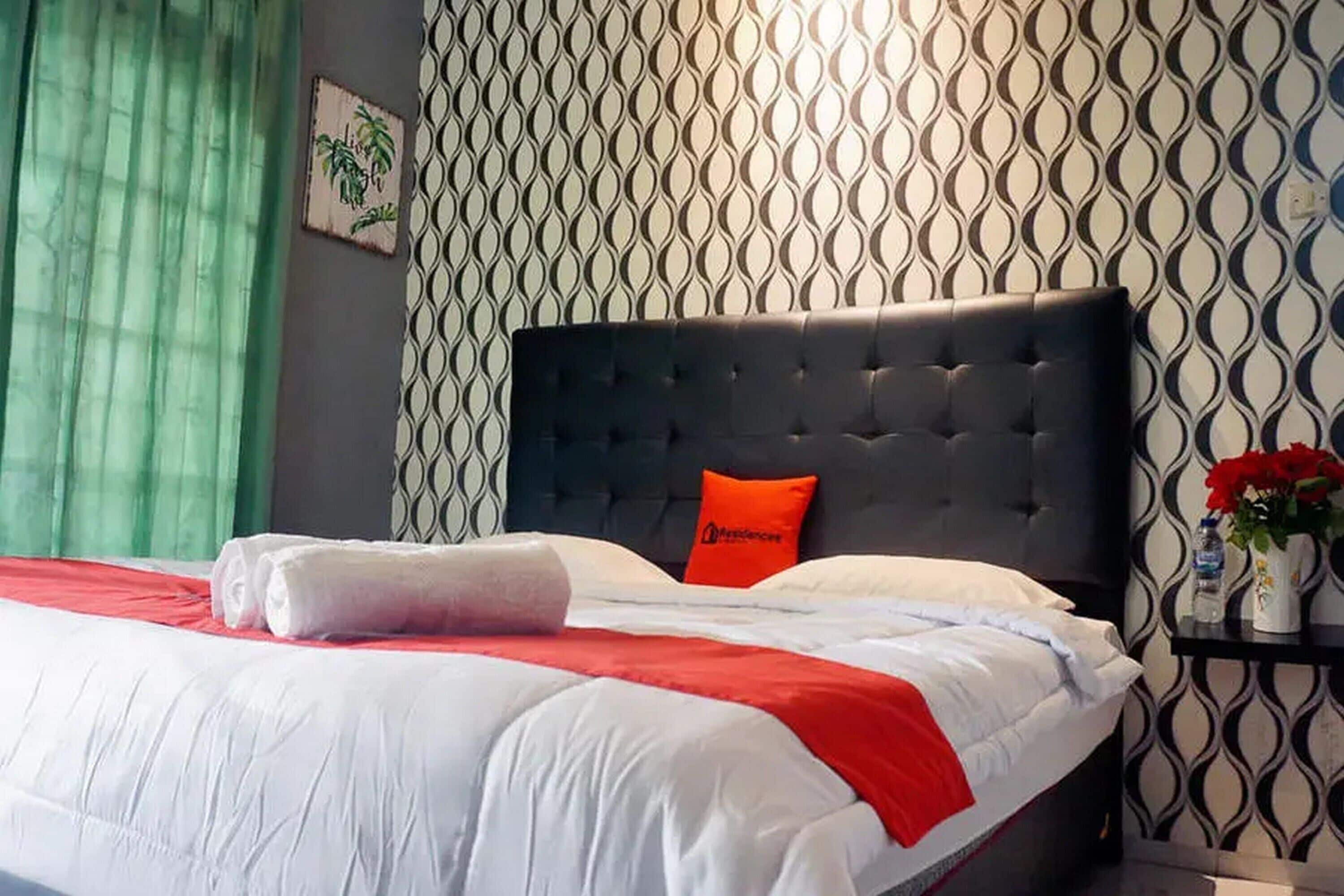 Un lit bien fait, avec une couvre-lit rouge et blanche, une tête de lit noire et un seul oreiller orange.