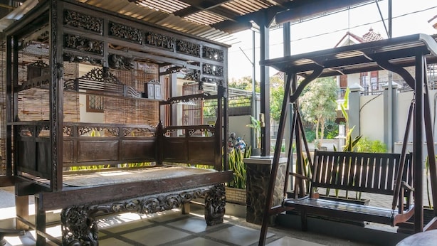 Un banco de madera tradicional con tallas intrincadas, una maceta de piedra y un sillón mecededor en un espacio al aire libre cubierto con suelo de baldosas.