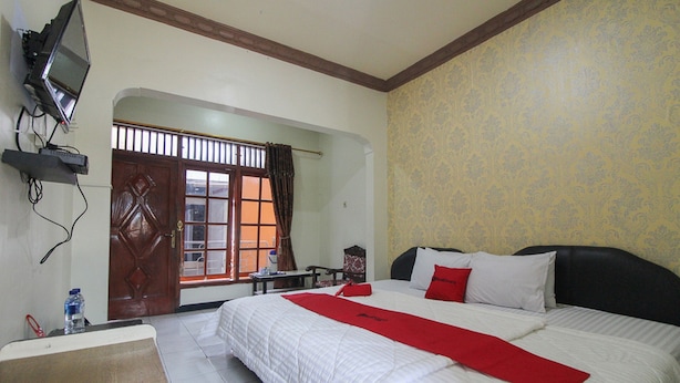 A hotel room with a bed, a TV mounted on the wall, a window with curtains, and a door leading to another room. - RedDoorz near Sarangan Lake (Plaosan)