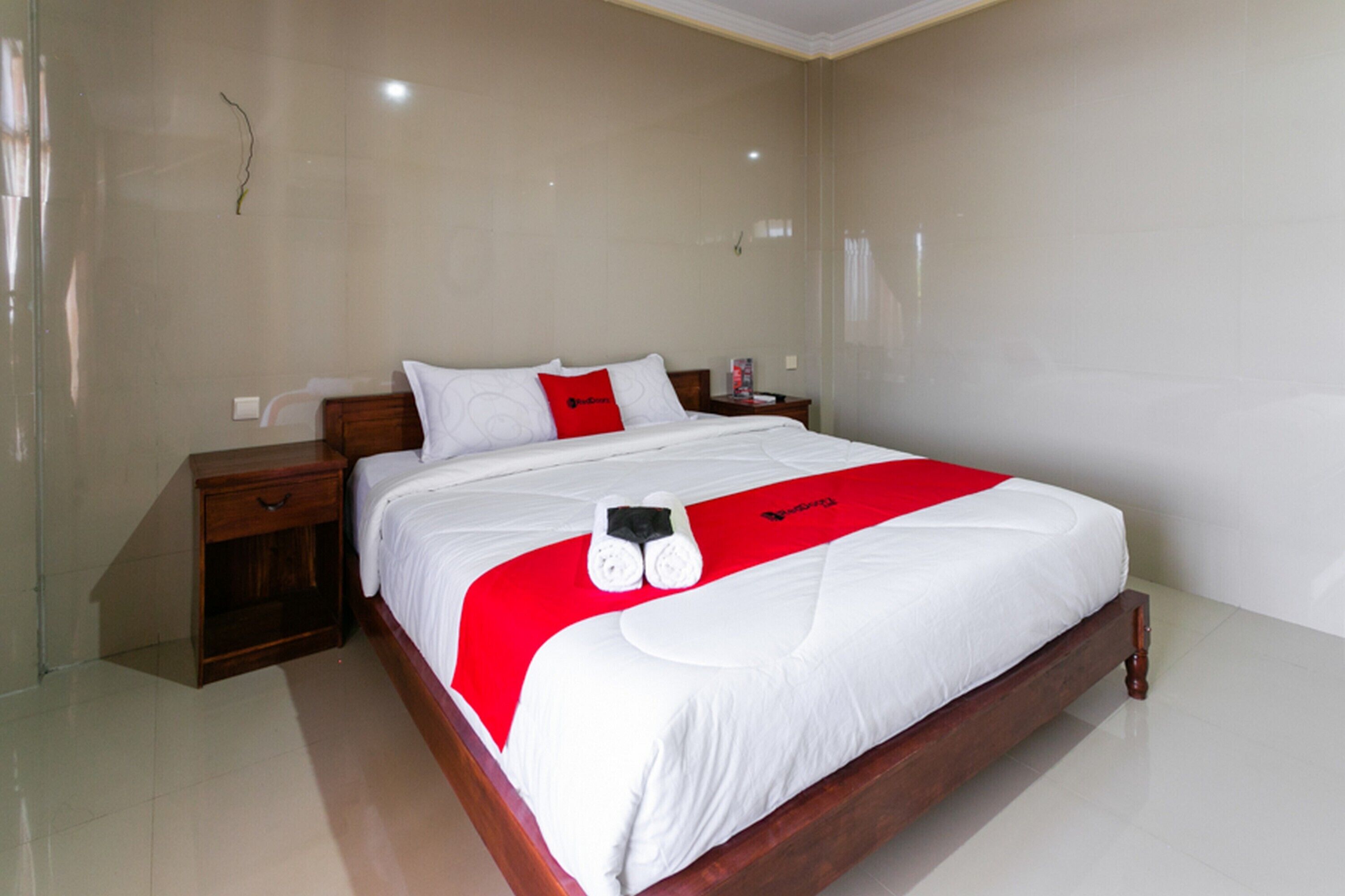 A neatly made bed with white bedding and a red accent pillow, a wooden nightstand, and a small bedside table with a lamp.