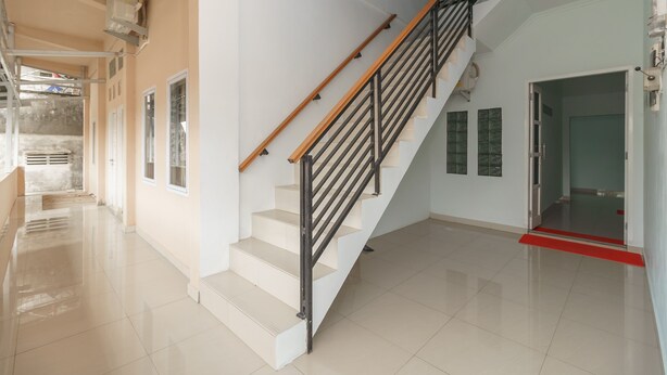 Una escalera con barandilla de madera y balaustradas de metal negro, que conduce a una habitación con suelo de baldosas y una puerta con felpudo rojo.