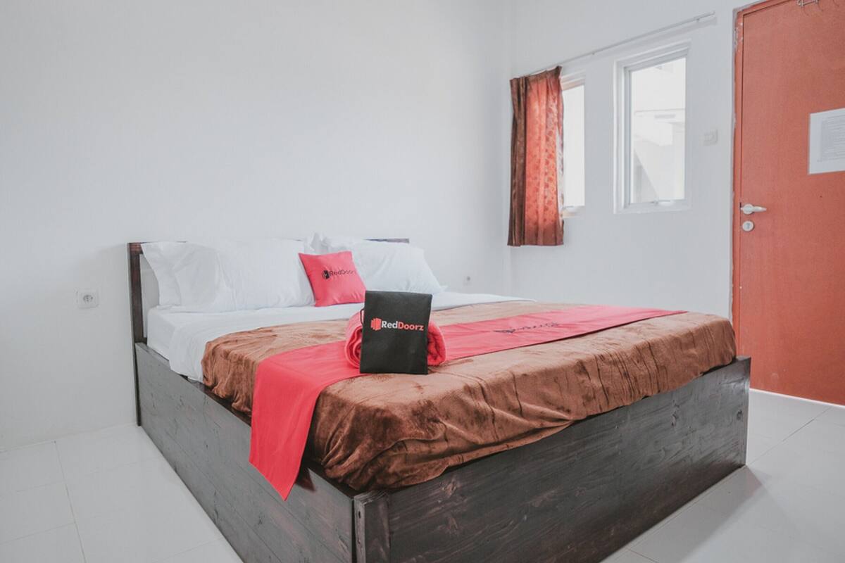 Ein Einzelbett mit braunem Kopfteil, weißer Bettwäsche und einer roten Decke. Eine schwarze Tasche mit dem Schriftzug "RedDoorz" liegt auf dem Bett.