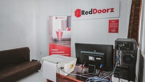 Een kamer met een bruine bank, een bureau met een computer en printer, een RedDoorz bordje en een banner die accommodatie aanbiedt.
