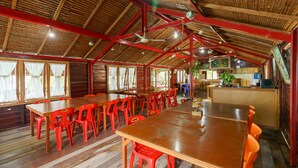 Un vaste espace dédié à la salle à manger, avec des tables en bois et des chaises en plastique rouge, un haut plafond en bois avec des poutres rouges, et de grandes fenêtres ornées de rideaux blancs.