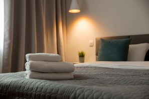 Ein ordentlich gemachtes Bett mit einer gemusterten Decke, ein Stapel gefalteter Handtücher und eine Nachttischlampe, die warmes Licht spendet.