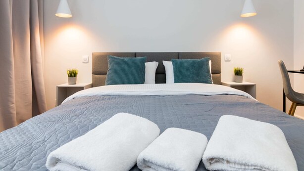 A neatly made bed with a grey quilt, white pillows, and two teal pillows. There are two bedside tables with small potted plants. - Jerozolimskie Central View Apartment (Warsaw)