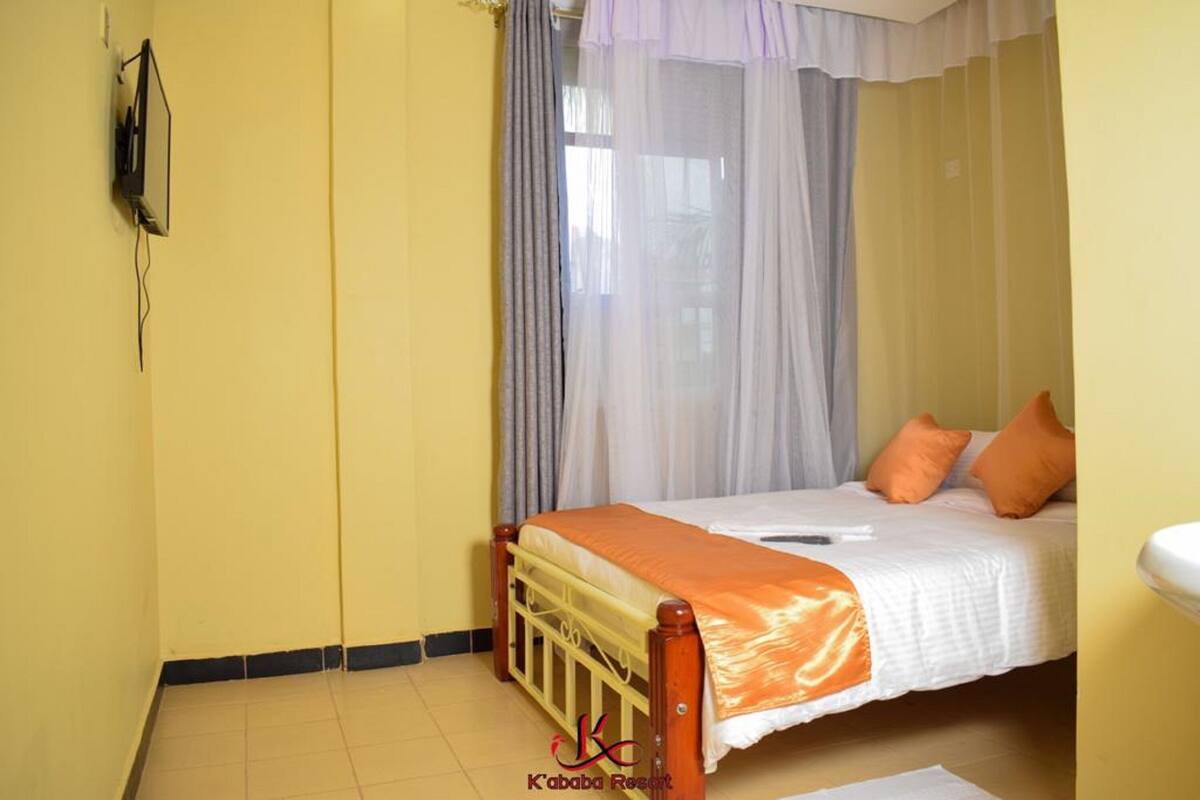 Una cama individual con una colcha naranja, cabecera de madera y dos almohadas naranjas. Un mosquitero cubre la ventana.