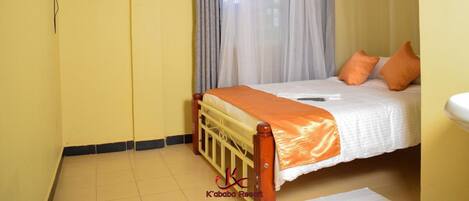 A single bed with an orange bedspread, a wooden headboard, and two orange pillows. A mosquito net is draped over the window.