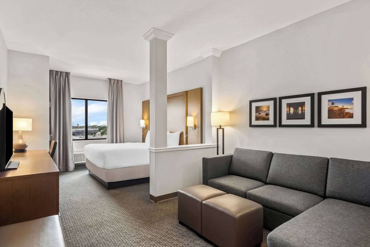 Habitación de hotel con cama, sofá, escritorio, televisor, lámpara y ventana con cortinas.