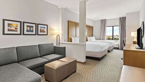 Habitación de hotel con un sofá gris, un pequeño reposapiés, una cama con ropa blanca, un cabecero de madera y un televisor sobre un escritorio de madera.