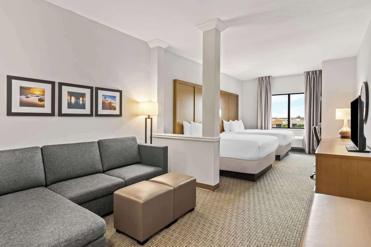 Habitación de hotel con un sofá gris, un pequeño reposapiés, una cama con ropa blanca, un cabecero de madera y un televisor sobre un escritorio de madera.