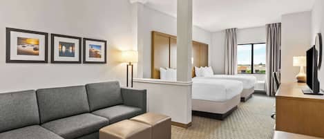A hotel room with a gray sofa, a small ottoman, a bed with white linens, a wooden headboard, and a television on a wooden desk.