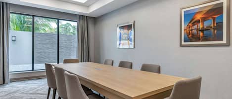 Een vergaderruimte met een lange tafel, zes stoelen, een plafondlamp, een groot raam met gordijnen, en twee ingelijste foto's aan de muur.