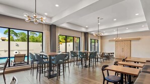 Un comedor interior moderno con varias mesas y sillas, ventanas amplias que ofrecen vistas a una piscina y follaje, e iluminación empotrada en el techo.