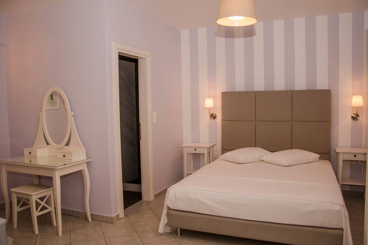 A bedroom with a bed, a bedside table, a nightstand, a dresser with a mirror, and a door.