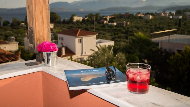Een boek met de titel 'Návn - Mani' op een balkon, met een glas rode drank en een metalen bekertje met roze bloemen.