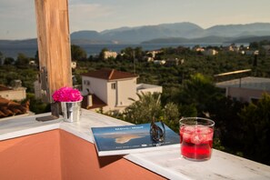 Un livre intitulé « Návn - Mani » est posé sur un balcon, à côté d’un verre contenant une boisson rouge et d’une tasse en métal ornée de fleurs roses.