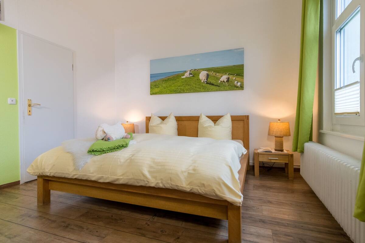 Quarto com cama de madeira, mesa de cabeceira com luminária, pintura de ovelhas na parede e vista para o mar pela janela.