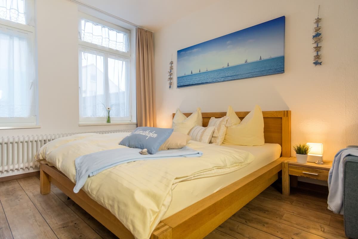 Quarto com cama de madeira, mesa de cabeceira com abajur, janela com cortinas e uma pintura de uma paisagem marítima.