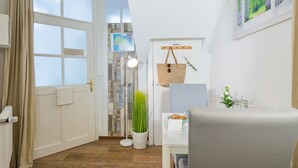 Une pièce avec un plancher en bois, une porte blanche portant l’inscription « Jardin », une suspension florale, une table avec une plante et une fenêtre donnant sur une forêt.