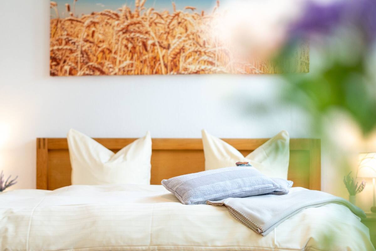 Cama bem arrumada com roupa de cama e almofadas brancas, cabeceira de madeira e um quadro de trigo na parede.