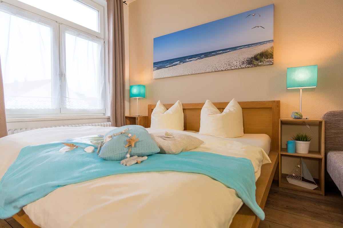 Cama bem arrumada com manta e travesseiros azuis, cabeceira de madeira, quadro de uma praia na parede e luminárias de cabeceira.