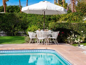 A poolside area with a white umbrella, white metal chairs, and a table with decorative items.