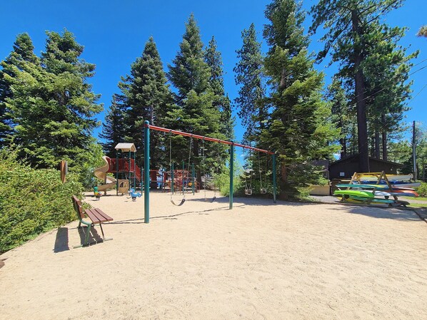 Un parque infantil con columpios y un tobogán, rodeado de árboles y un suelo de arena.