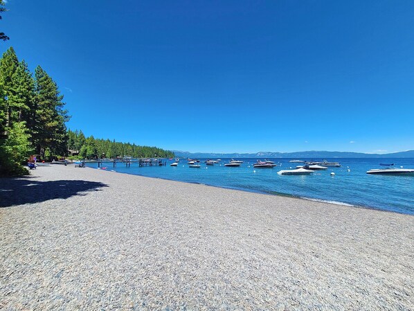 A pebbled beach with boats on calm water, trees, and a clear sky. - Stine Haus - 3 Bedroom, 2.5 Bath - HOT TUB & Beach Access! (Tahoe City)
