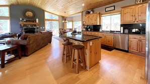 Una cocina amplia con armarios de madera, una isla central con taburetes, una chimenea de piedra y ventanas grandes.
