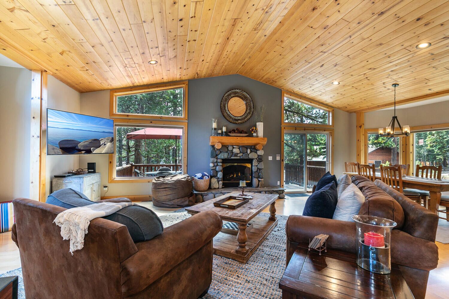A cozy living room with a fireplace, wooden ceiling, and large windows.
