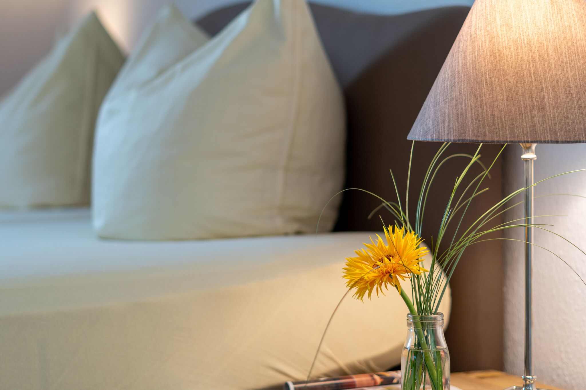 Ein Bett mit weißen und braunen Kissen, ein Nachttisch mit einer Lampe und einer Vase mit gelben Blumen.