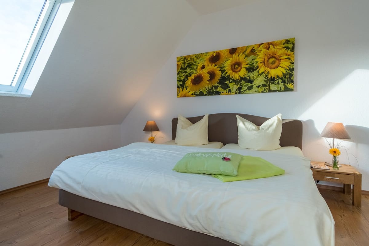 A bedroom with a large bed, two bedside tables with lamps, a painting of sunflowers on the wall, and a skylight.