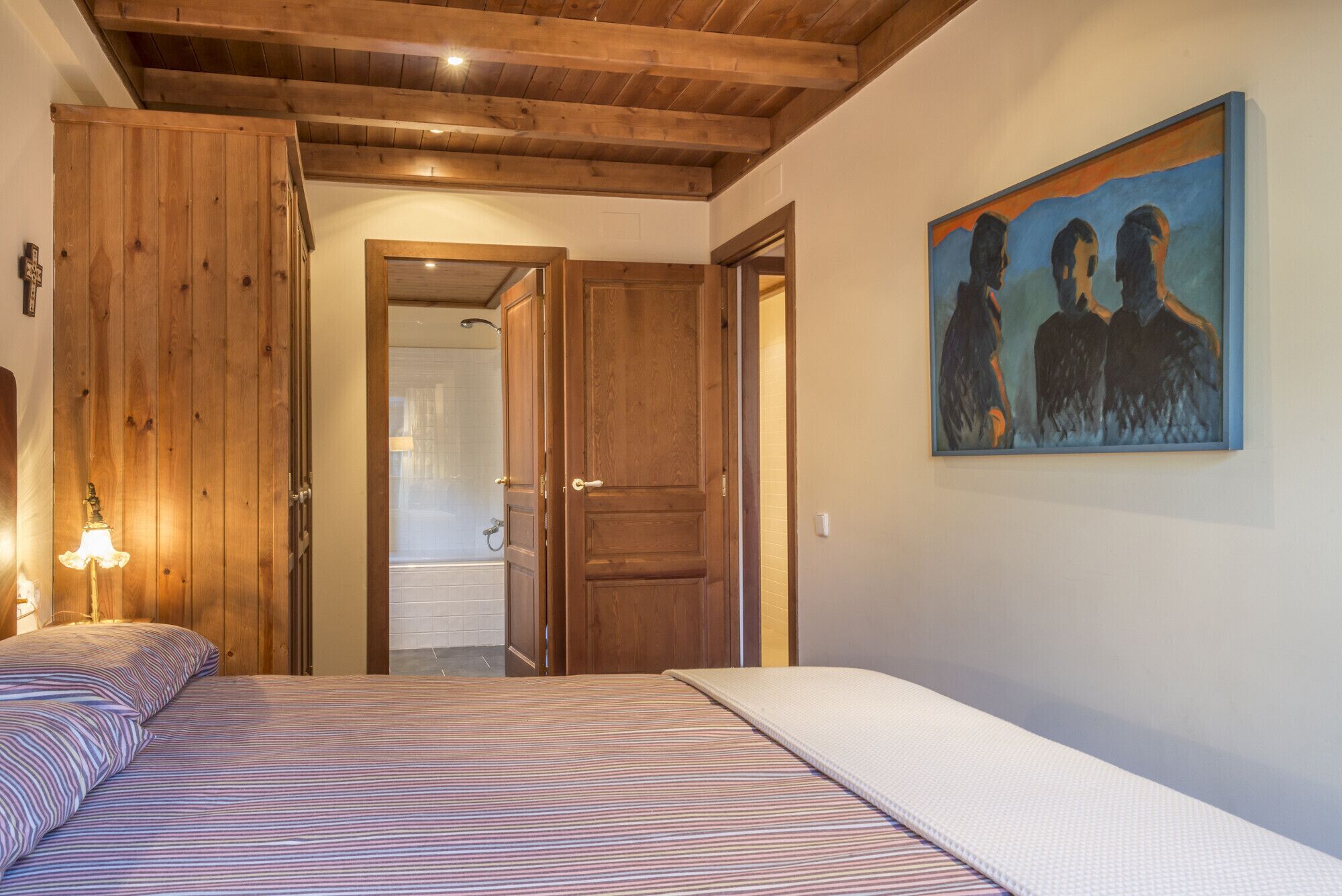 Un dormitorio con cama, techo de madera, un cuadro y baño visible a través de una puerta abierta.