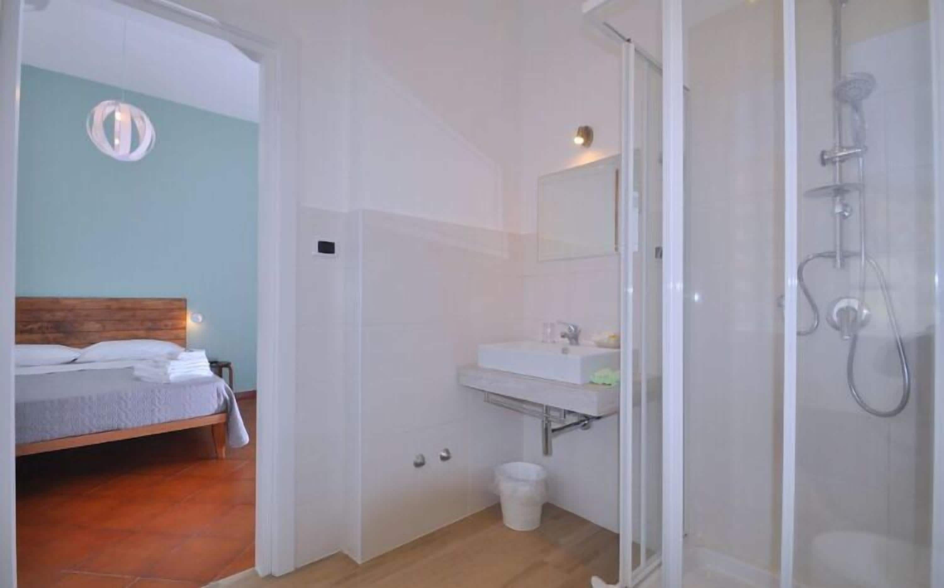 Une chambre à coucher avec un lit doté d’une tête de lit en bois, une petite table de chevet et une commode. À côté de la chambre se trouve une salle de bain avec une douche à parois de verre, un lavabo blanc et un miroir au-dessus.