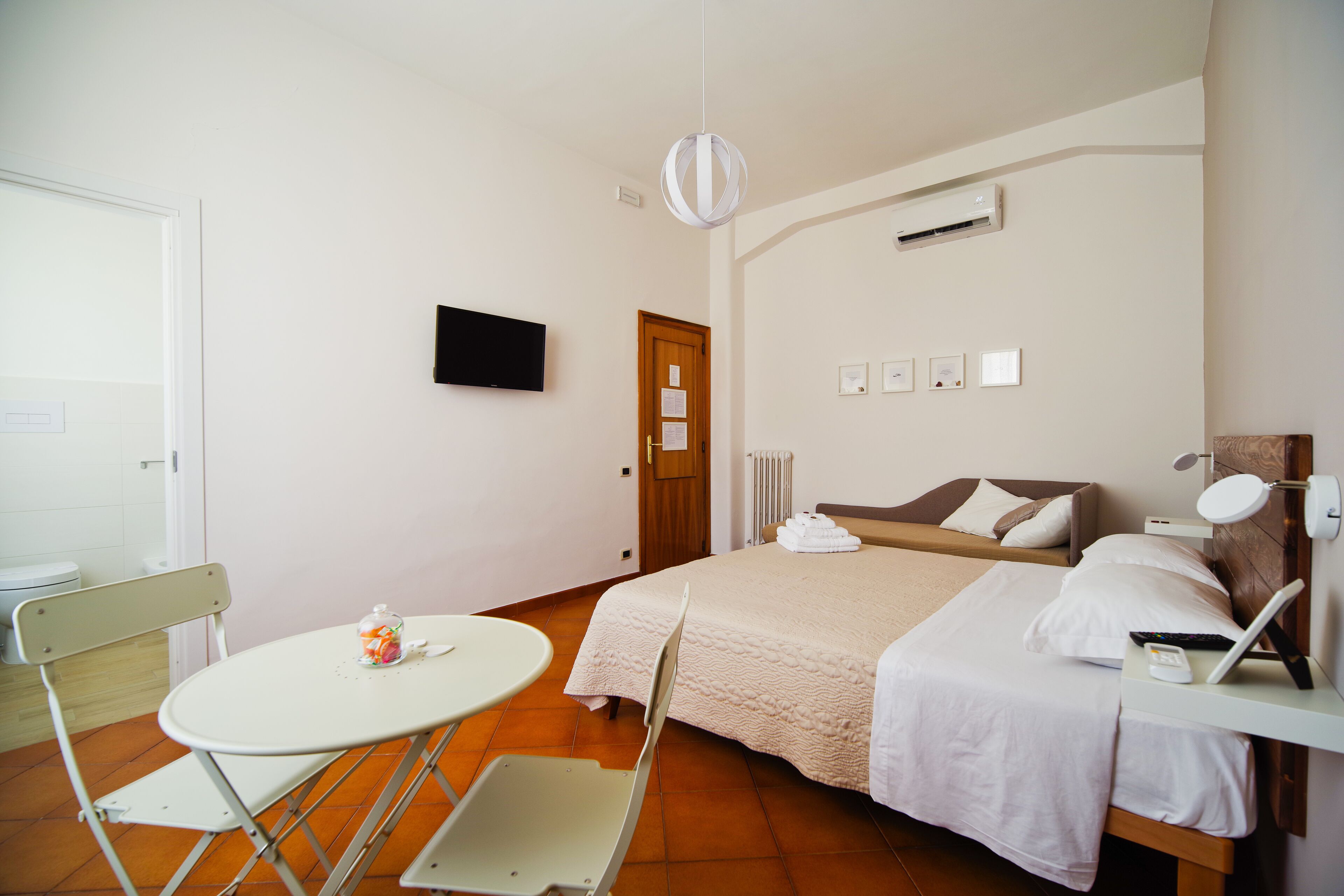 A hotel room with a bed, bedside table, chair, and a small table with a bottle and glass.