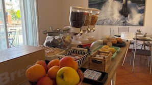 A breakfast buffet with various food items, a coffee dispenser, and a view of the sea through a window. - B&B Il Faro (Salerno)