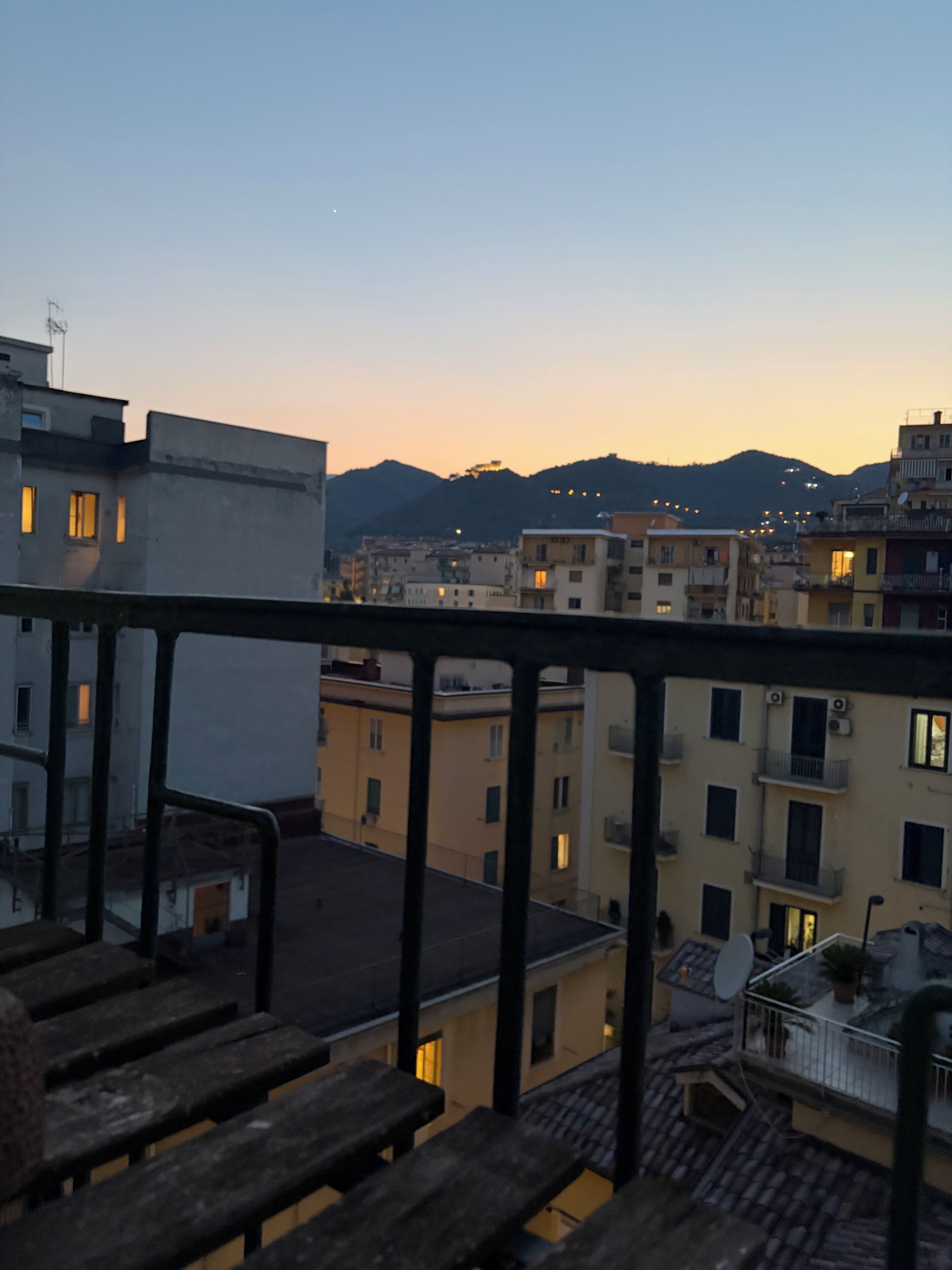 Un paisaje urbano al anochecer con edificios, una montaña al fondo y un balcón con barandillas de madera.