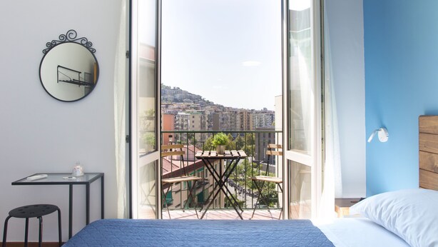 A bedroom with a bed, a small table with a chair, a round mirror, and a balcony with a view of buildings. - B&B Il Faro (Salerno)