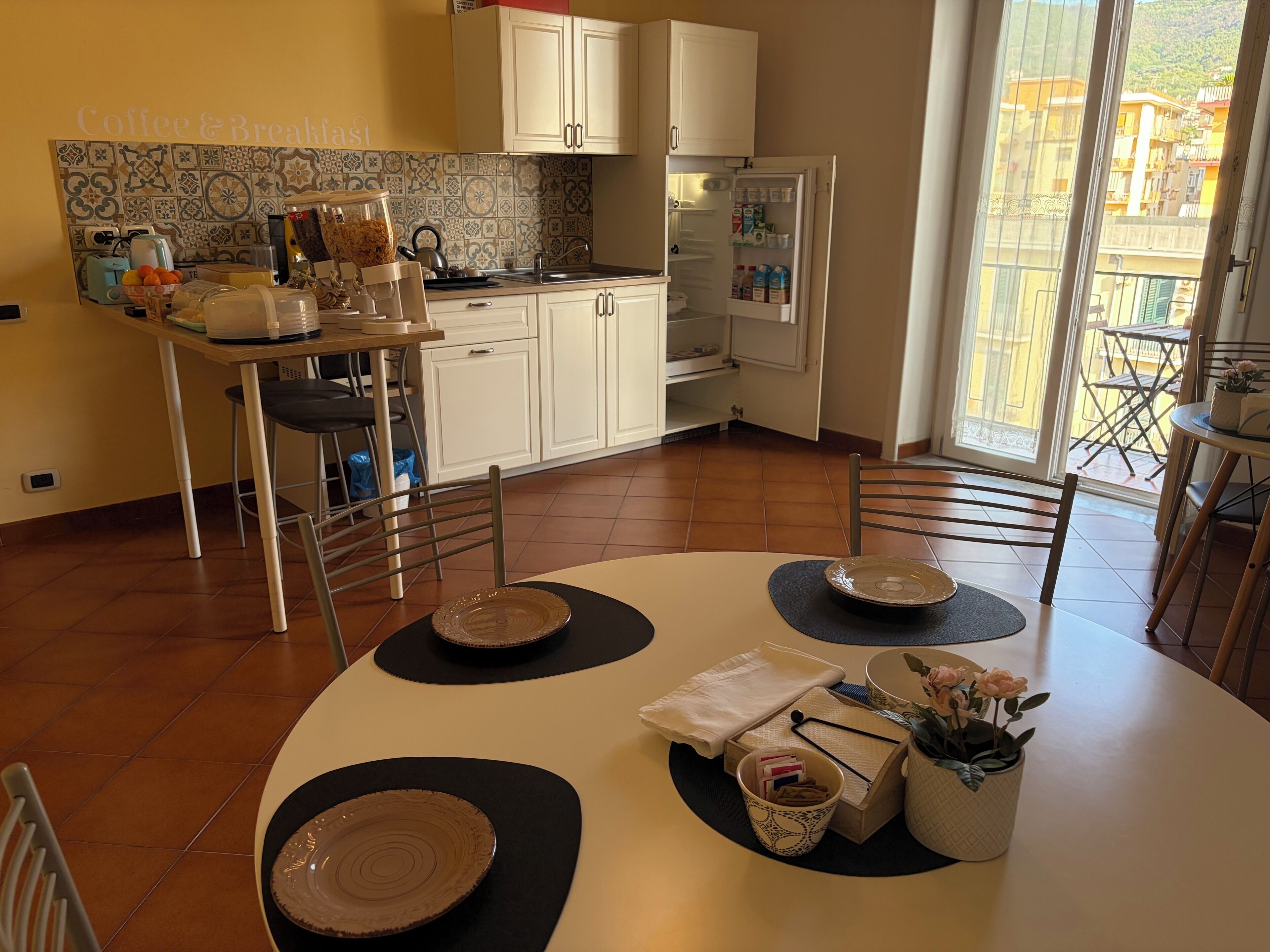 A kitchen with white cabinets, a tiled backsplash, and a refrigerator. There is a dining table with plates and a vase with flowers.