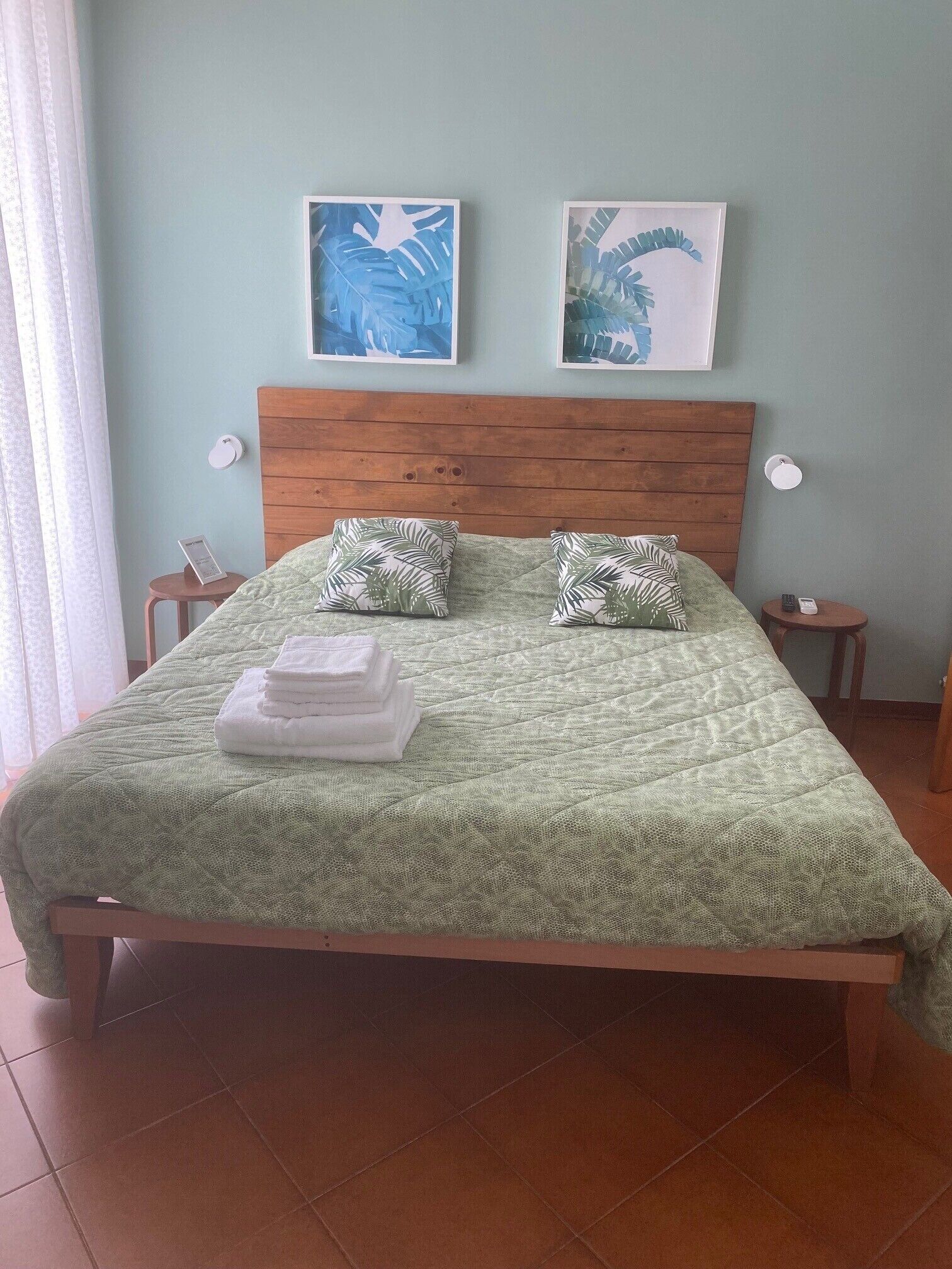 Uma cama bem arrumada com um edredom verde, duas almofadas estampadas com folhas e dois quadros na parede.