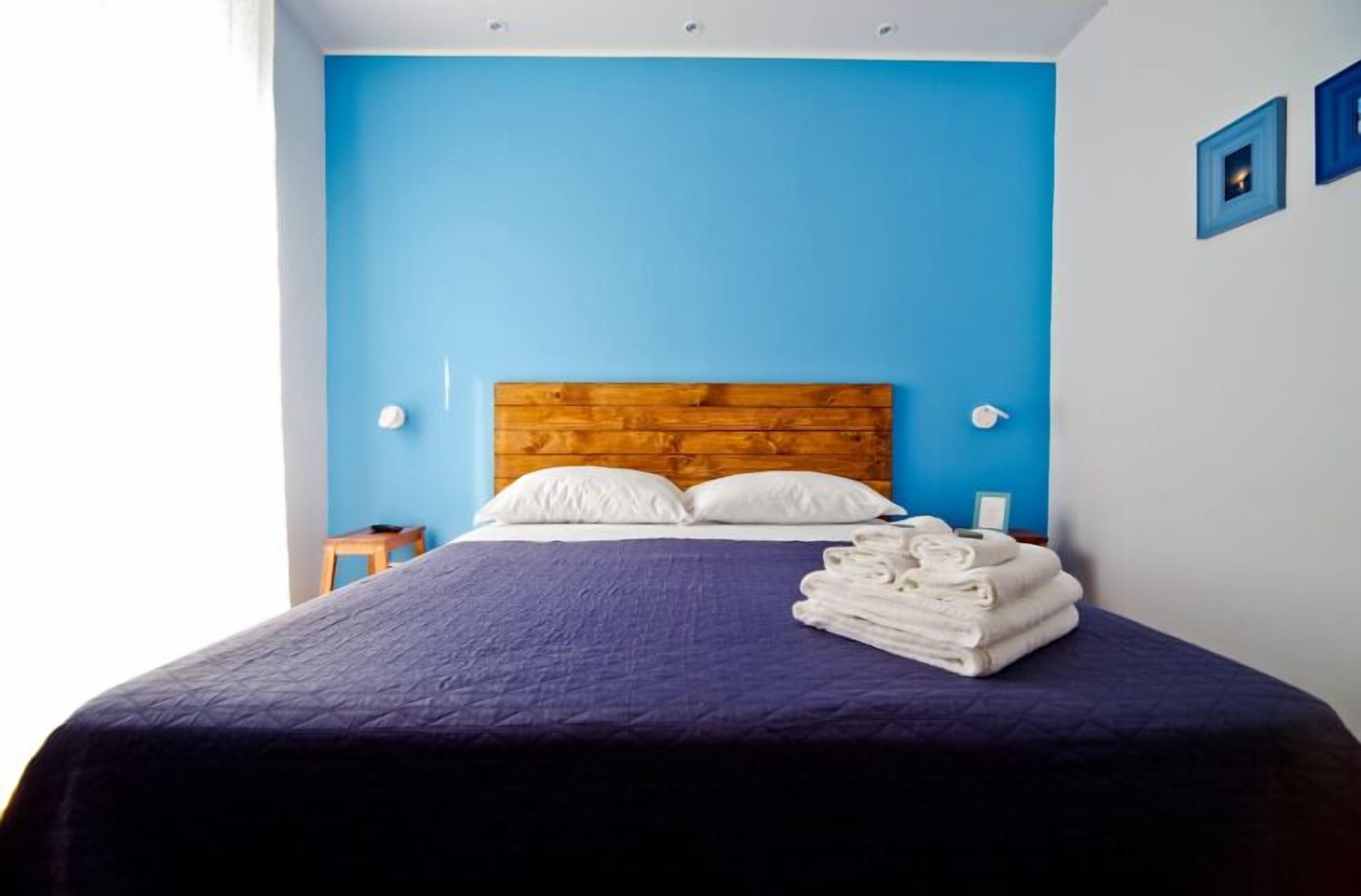 A bedroom with a blue wall, wooden headboard, white pillows, and a neatly folded stack of towels on the bed.