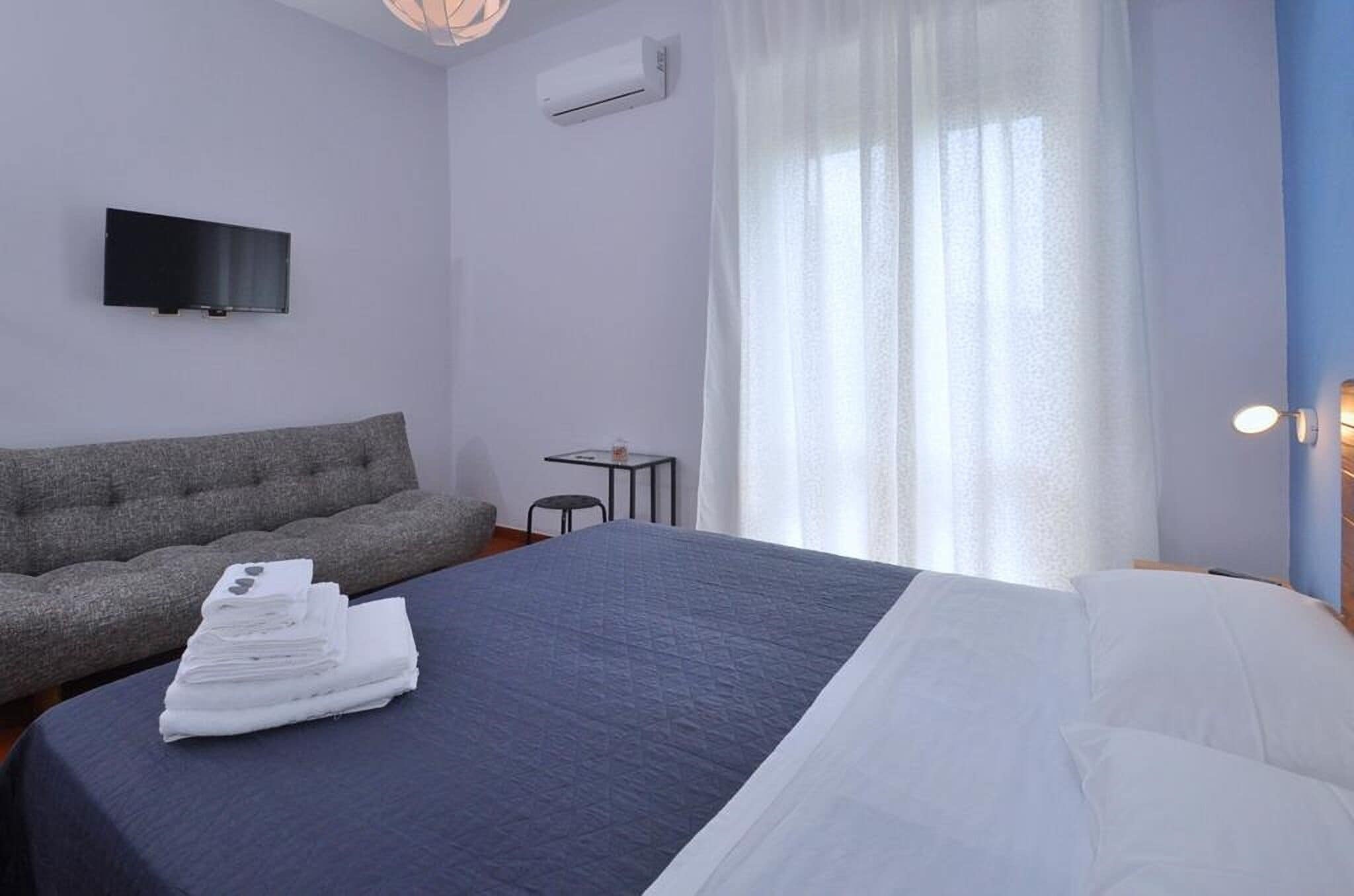 A hotel room with a bed, a sofa, a TV, and an air conditioning unit.