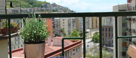 A balcony with a potted plant and a decorative bowl, overlooking a cityscape with buildings and greenery.